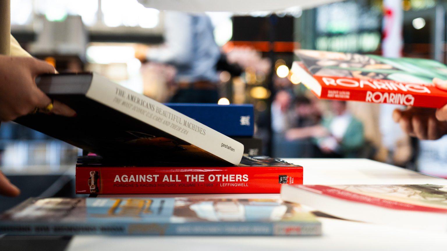 Hände, die Bücher über einem Tisch stapeln oder austauschen, mit einem roten Buch im Vordergrund mit dem Titel 'AGAINST ALL THE OTHERS' und unscharfen Messebesuchern und Lichtern im Hintergrund.